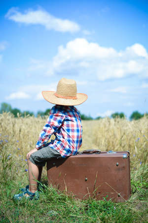Picture of little boy in cowboy hat sitting on retro suitcase in countryside. Backview of kid beside wheat field on blue sky sunny outdoors background.の写真素材