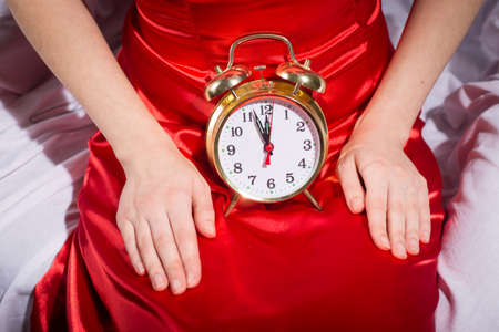 Picture of vintage alarm clock on woman's knees. Closeup of woman in red evening dress sitting on bed at almost midnight on bedroom indoor background.の写真素材