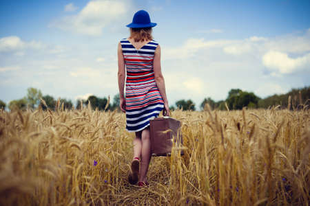 Picture of elegant female in blue hat with retro suitcase walking in wheat field. Backview of girl in striped dress on blue sky summer countryside background.の写真素材