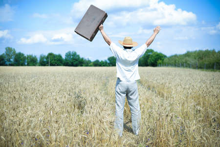 Picture of happy excited man wearing straw hat with suitcase rising hands in wheat field. Backview of male in white shirt excited on golden countryside background.の写真素材