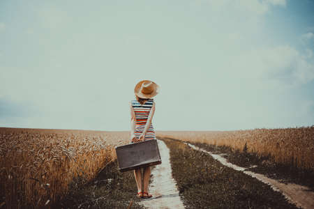 Picture of young woman with old suitcase standing on road between wheat field. Backview of girl in stripen dress and straw hat on sunless countryside background.の写真素材