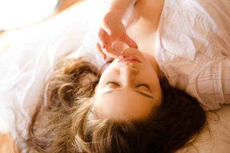 Picture of beautiful girl dreaming with closed eyes in morning sunlight. Charming woman touching her lips gently on sunny indoor background.の写真素材