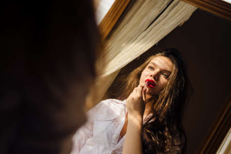 Picture of young beautiful woman in white shirt applying red lipstick. Mirroring image of pretty girl with tangled hair on countryhouse indoor background.の写真素材