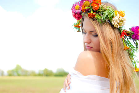 Portrait of charming beautiful girl in flower wreath looking down. Young lady in white chiffon on blurred light summer outdoor background.の写真素材