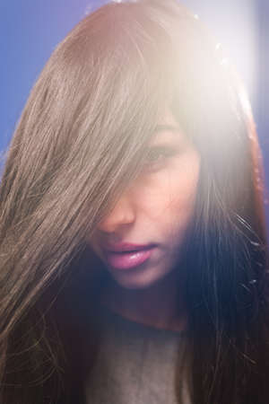 Portrait of beautiful young lady looking from under long hair. Touching teen girl on lens flare blue...