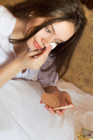 Portrait of charming girl lying on bed and applying powder on her face. Young woman with long hair smiling on blurred indoor background.の写真素材