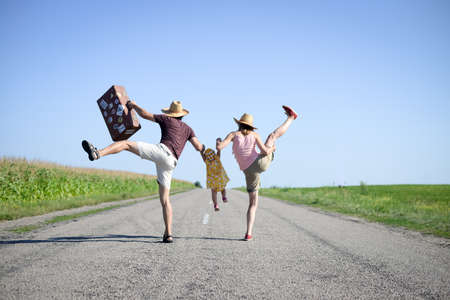 Picture of merry family jumping and dancing on road in summertime. Backview of parents and baby girl with retro suitcase on sunny sky countryside background.の写真素材
