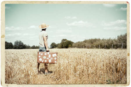 Picture of man in straw hat with old suitcase standing on wheat field. Framed image in vintage paper photo style.の写真素材