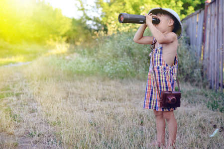 Picture of boy wearing plaid romper looking in spyglass near old wooden fence. Little explorer with camerabag on sunny countryside background.の写真素材