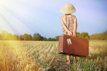 Picture of little boy wearing pith helmet carrying old suitcase in countryside. Backview of kid in plaid romper walking away on sunny flare background.の写真素材