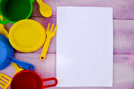 Picture of wooden tabletop with colorful toy dishware and blank sheet of paper. Plastic pates and cutlery on lilac rustic with copy space background.の写真素材