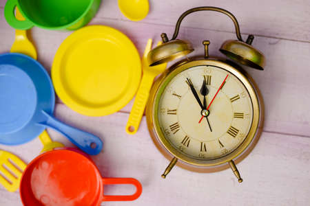 Picture of wooden tabletop with colorful toy dishware and vintage alarm clock. Plastic pates and cutlery with old clock on lilac plank background.の写真素材