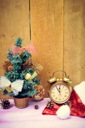 Picture of little christmas tree decorated with bows and old alarm clock. Artificial tree, pine cones and santa hat on wooden indoor background.の写真素材