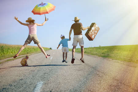 Picture of amazing happy family jumping on country road in summer. Backview of parents and two kids with suitcase and rainbow umbrella on sunny countryside background.の写真素材