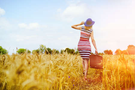 Picture of young woman in blue hat with suitcase walking away through golden wheat field. Backview of girl in striped dress in direct sunlight on summer countryside background.の写真素材