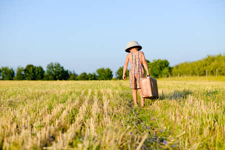 Picture of little boy wearing plaid romper and pith helmet carrying big suitcase in grass field over blue sky sunny outdoors background. Kid in safari helmet walking away.の写真素材