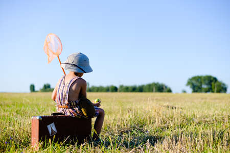 Picture of sweet small boy sitting on old brown suitcase eating green apple. Kid has ring net and flask over blue sky sunny outdoors backgroundの写真素材