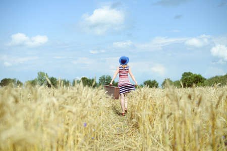 Female wearing hat with suitcase walking away through wheat field. Backview of girl in striped dress...