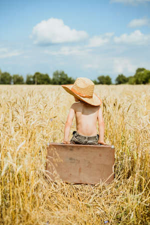 Little kid sitting on big old brown leather suitcase in summer countryside. Child with valize wearing straw hat in wheat field over sunny blue sky outdoors backgroundの写真素材
