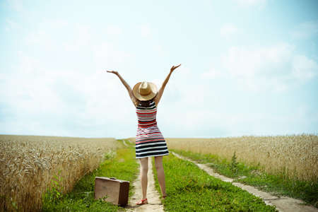 Backview of happy woman with old suitcase on countryside road. Girl with valize wearing hat and striped dress rising hands to the sunny blue sky backgroundの写真素材