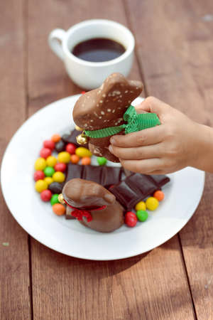 Picture of child hand holding big chocolate bunny. Plate with colorful candies and cup of coffee on rustic plank table background.の写真素材