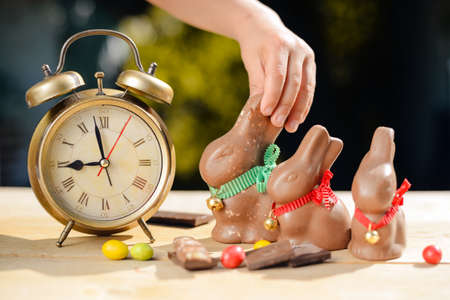 Picture of child hand taking big chocolate bunny. Three bunnies and candies beside retro alarm clock on table top blurred background.の写真素材