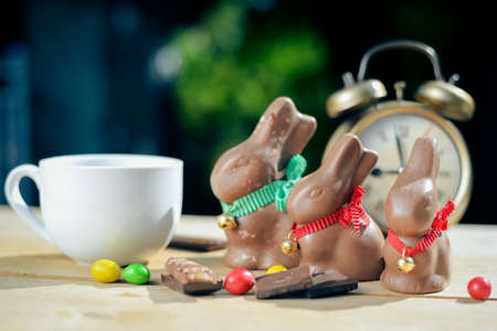 Picture of three big chocolate bunnies on table top. Colorful candies  and white cup beside retro alarm clock on blurred indoor background.の写真素材