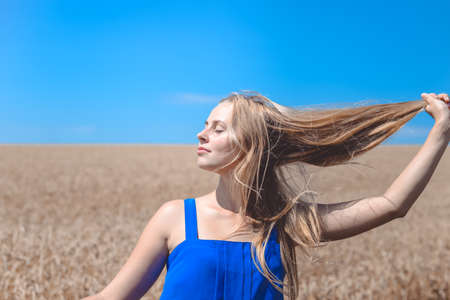 Portrait of pretty girl holding her long hair with closed eyes. Young woman dreaming on blue sky and golden wheat field background.の写真素材