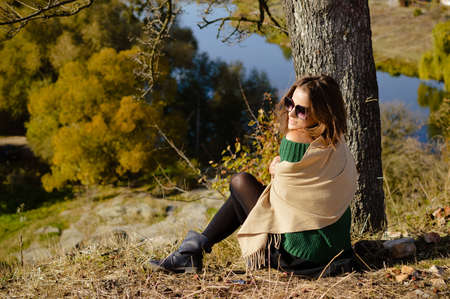 Picture of young beautiful woman in shawl and sunglasses on riverside. Pretty girl sitting under old tree on sunny autumn outside background.の写真素材
