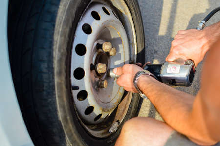 Picture of man beside car changing old wheel. Closeup of hands working with professional screwdriver on blurred sunny outdoor background.の写真素材