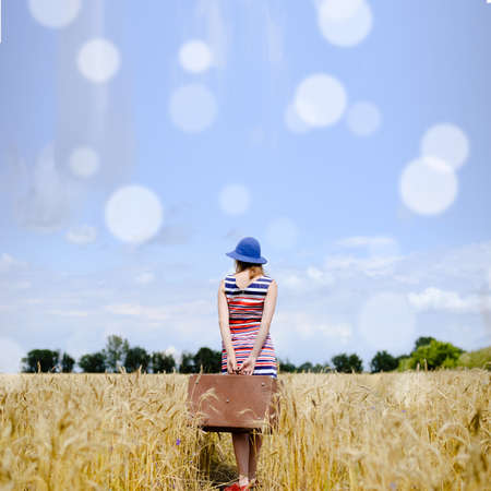 Picture of pretty girl in blue hat holding old valize. Young woman in striped dress standing in wheat field on lens flare sunny sky background.の写真素材