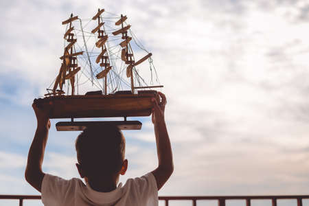 Picture of little boy holding beautiful ship model. Silhouette of happy kid on sunset sky outdoor background.の写真素材