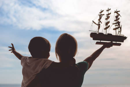 Picture of young woman and boy holding ship model. Silhouette of excited happy family on sunset sky outdoor background.の写真素材