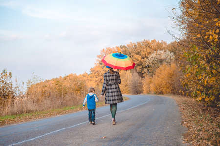 Picture of woman with rainbow umbrella walking with little boy on autumn countryside road. Backview of happy family on golden trees outdoor background.の写真素材
