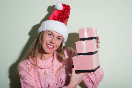 Portrait of joyful beautiful young lady in Santa hat holding three pink gift boxes. Pretty girl in pink sweater happy smiling on light indoor background.の写真素材