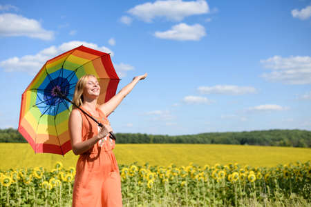 Portrait of joyful beautiful young lady in orange dress with rainbow umbrella smiling beside sunflowers field. Exciting girl with closed eyes on sunny sky countryside background.の写真素材