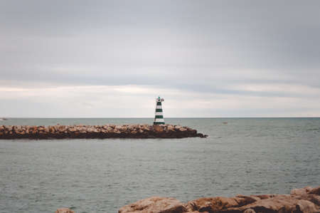 Picture of striped lighthouse on rocky pier in cold sea. Symbol of hope on sombre sky and grey water...