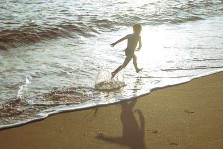 Picture of exciting little boy running on beach beside waves. Vintage image of kid in evening sunlights on seaside background.の写真素材