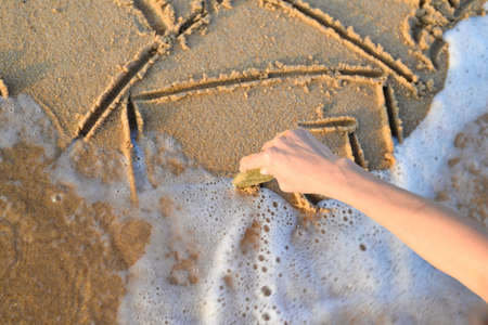 Picture of hand drawing house on sand. Closeup of sketching in surf on sunny seaside background.の写真素材