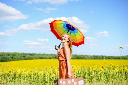Portrait of beautiful young lady with rainbow umbrella and retro suitcase beside sunflowers field. Pretty girl laughing on sunny countryside background.の写真素材