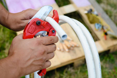 Picture of cutting metal-plastic pipe by special red scissors. Closeup of male hands working with white on pipe on blurred summer backyard background.の写真素材