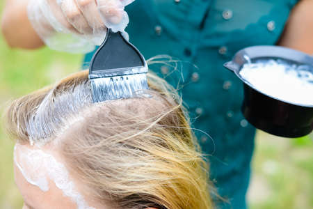 Picture of coiffeur coloring client's head with brush and bowl. Closeup of hands in gloves and blond hair on summer outdoor background.の写真素材