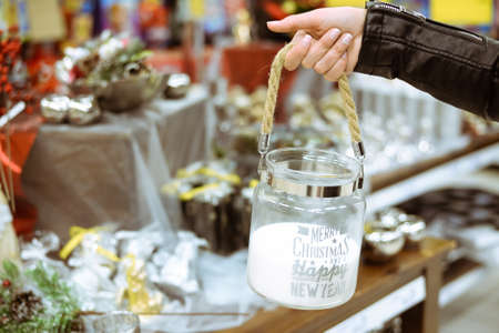 Picture of big white candle in glass box with rope handle. Closeup of hand holding christmas decor on blurred store indoor background.の写真素材