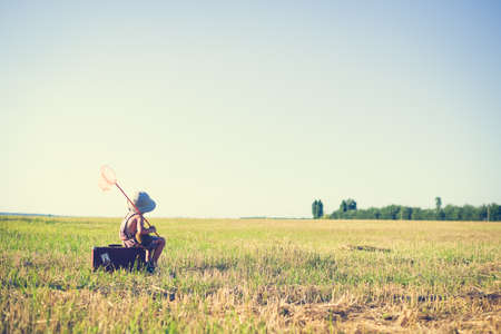 Picture of little boy sitting on old brown suitcase with butterfly net. Kid resting on sunny summer countryside background.の写真素材