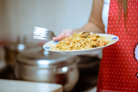 Picture of female hand holding plate with beautiful seafood pasta and forks. Tasty traditional dish on blurred kitchen indoor background.の写真素材