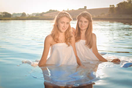 Picture of 2 beautiful princess young ladies in white dresses on summer sunset water outdoors backgroundの写真素材