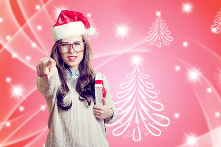 Picture of pointing finger and holding present box beautiful young lady in Santa Claus hat. Girl in glasses happy smiling and looking at camera on abstract festive design backgroundの写真素材