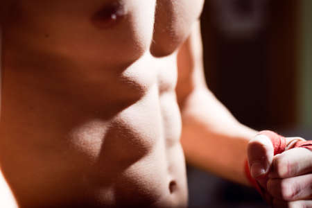 Young man training box punches with bandages closeup on strong muscles of torso over dark studio backgroundの写真素材