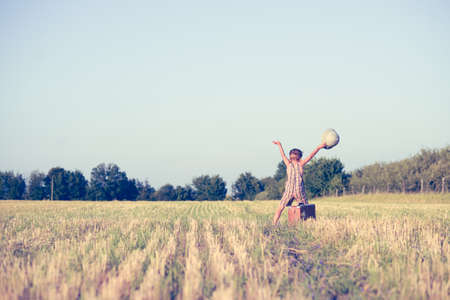 Picture of happy boy with raised hands in country field. Little explorer with pith helmet and old suitcase on blurred countryside background.の写真素材