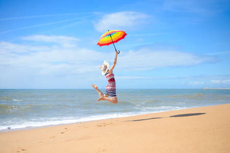 Picture of beautiful young lady holding rainbow umbrella and jumping on golden seashore. Backview of...